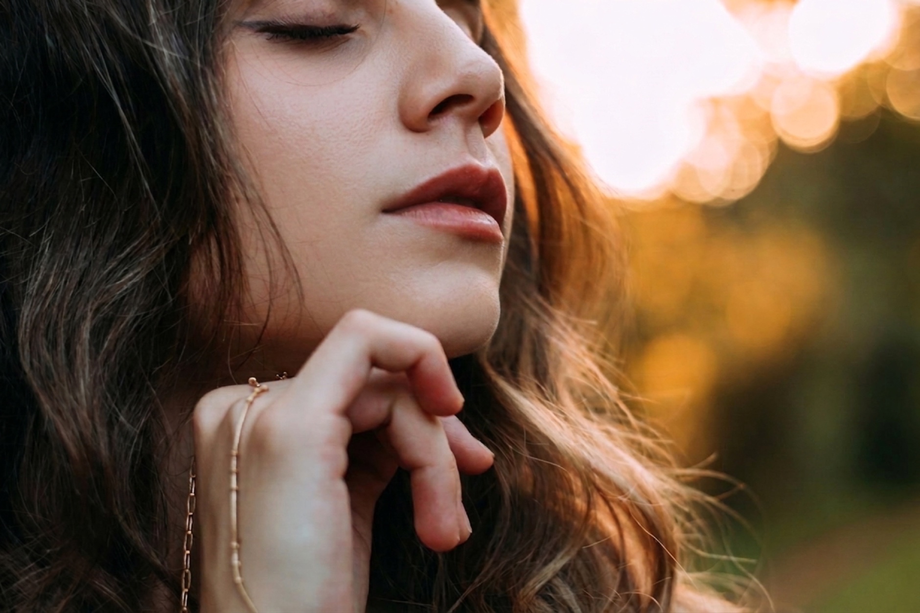 A woman with eyes closed in warm golden-hour light, glowing skin, soft bokeh behind her