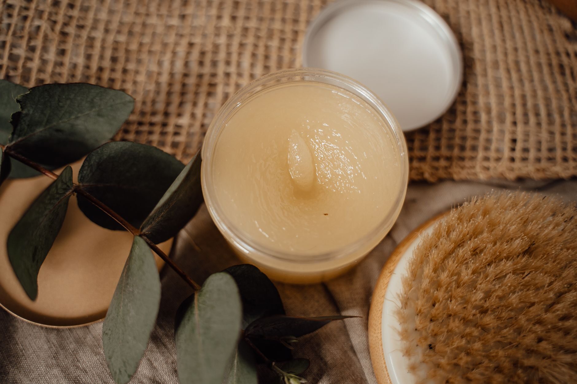 An open glass jar of golden scrub with eucalyptus leaves and a natural bristle brush