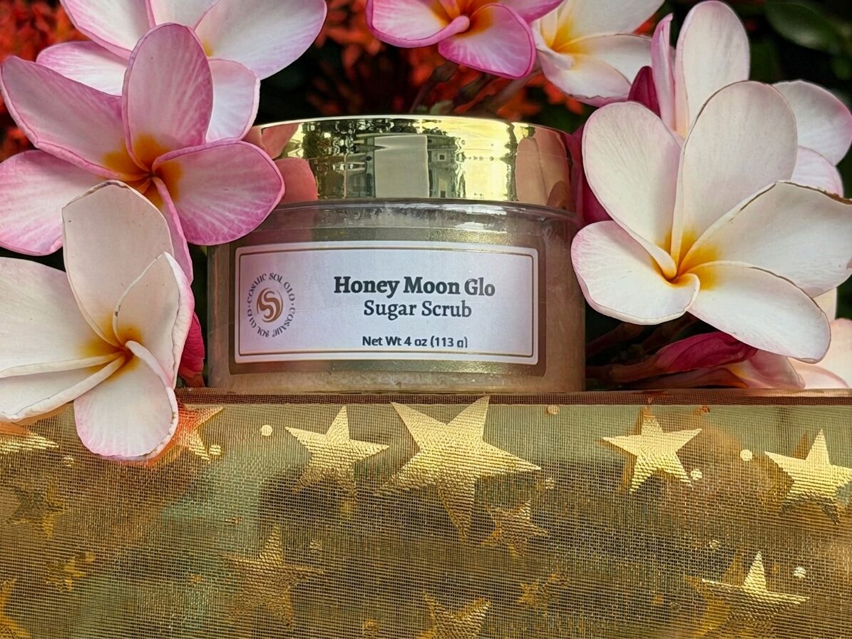 Honey Moon Glo sugar scrub jar with pale plumeria flowers over golden star fabric