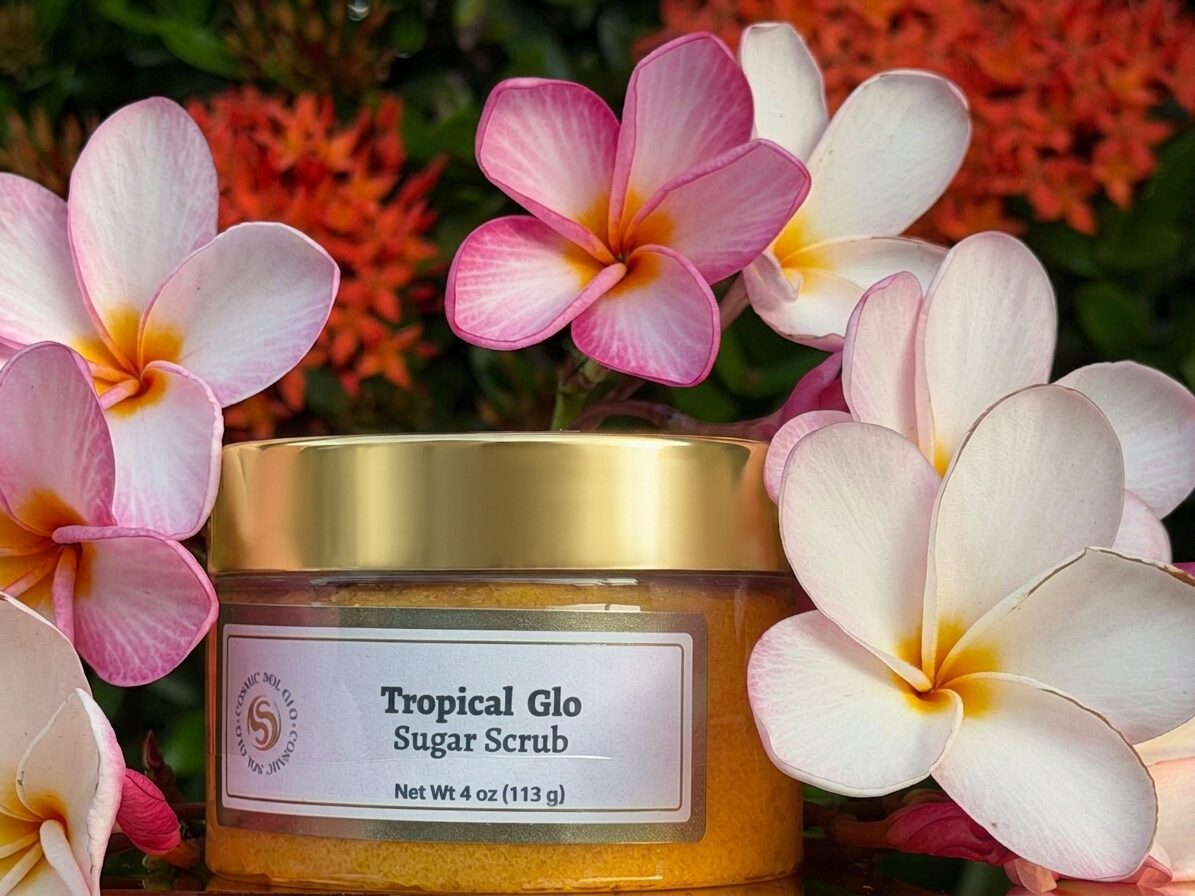 Tropical Glo sugar scrub in a gold-lid glass jar nestled in pink and coral plumeria